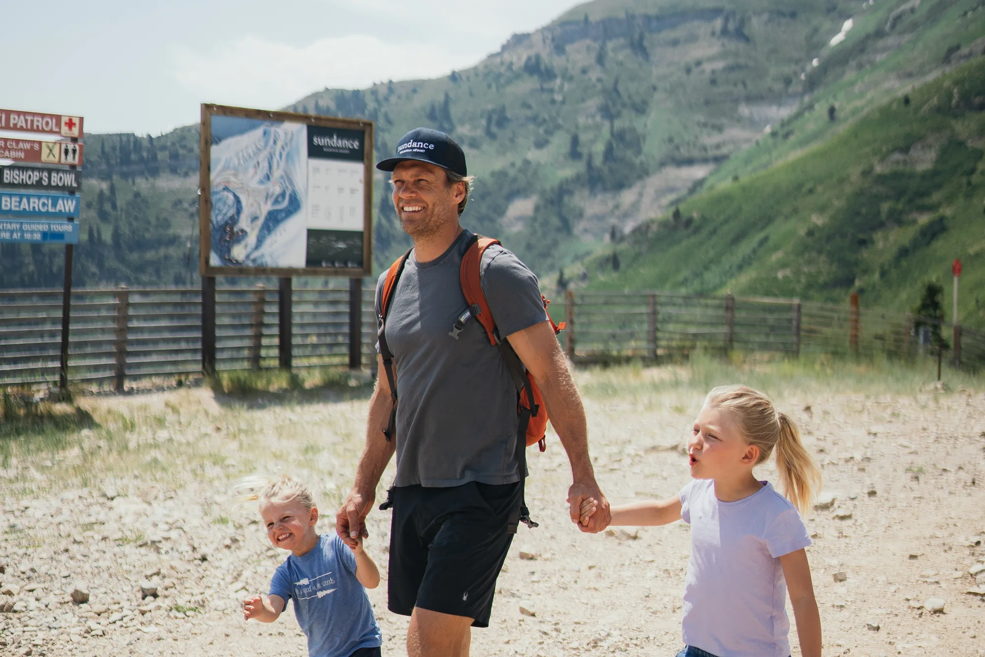 A man holding hands with two young girls walks on a rocky trail in a mountainous area, with green hills and blue sky in the background. All three are smiling and dressed in casual outdoor clothing.